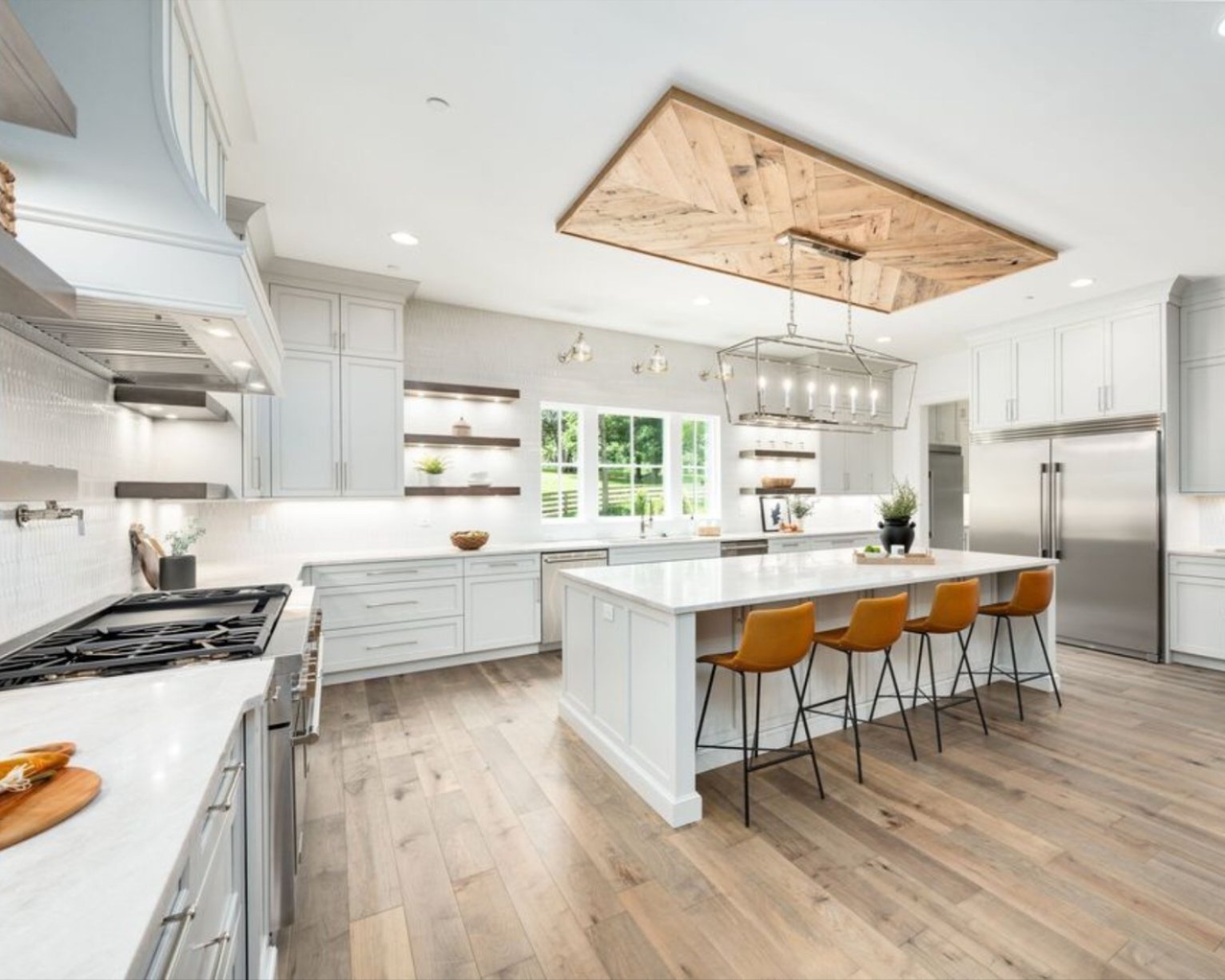 Modern open-concept kitchen featuring wide-plank light oak luxury vinyl flooring, soft gray shaker cabinets, white quartz countertops, a large center island with four caramel-colored bar stools, stainless steel appliances, floating wood shelves, oversized