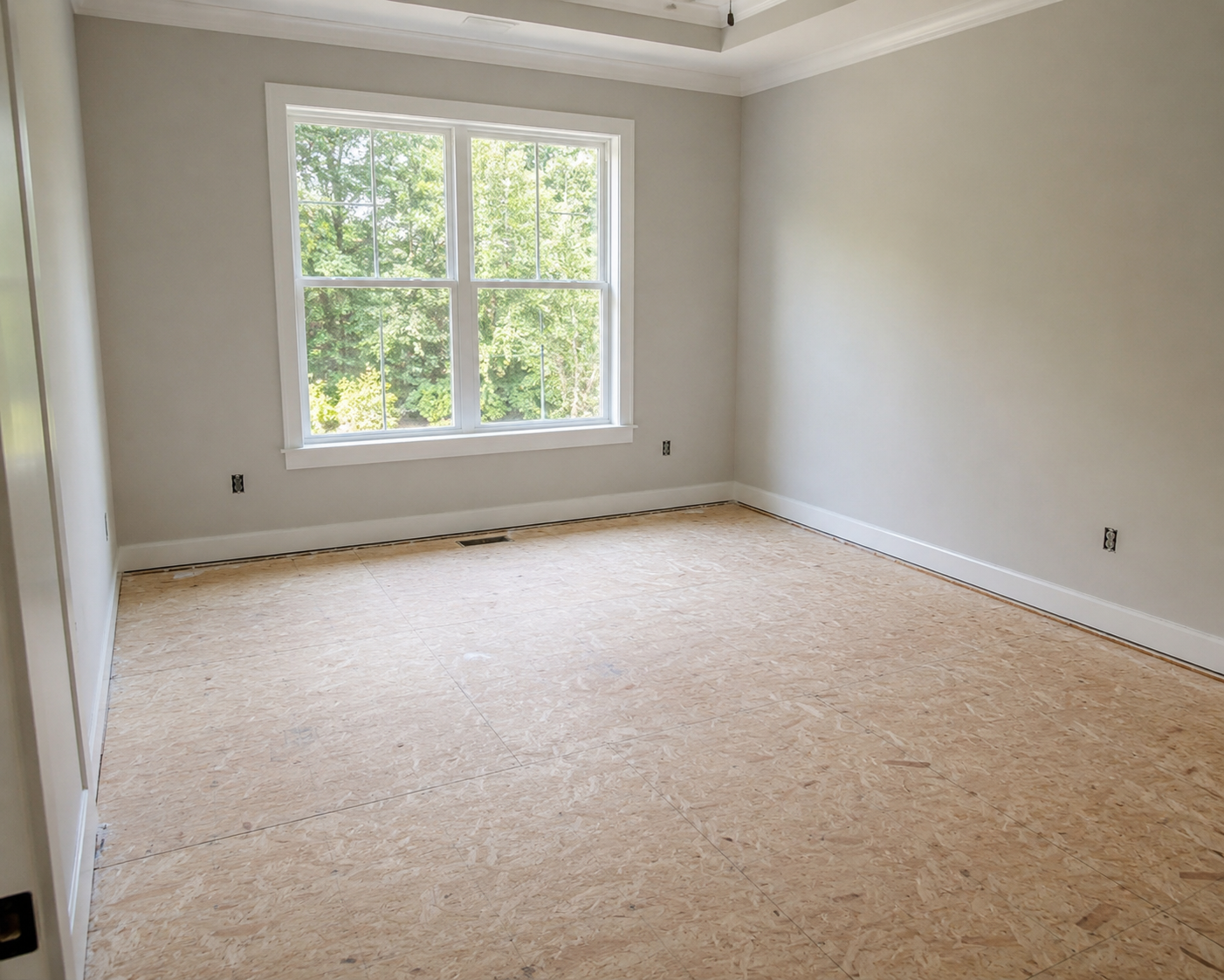 Empty bedroom prepared for new flooring installation, featuring an exposed wood subfloor, neutral walls, white trim, and a large window overlooking green trees.